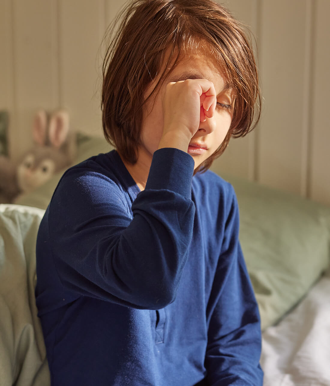 En dreng i blå pyjamas sidder på kanten af sin seng og kløer sit øje, som måske generer ham på grund af husstøvmideallergi