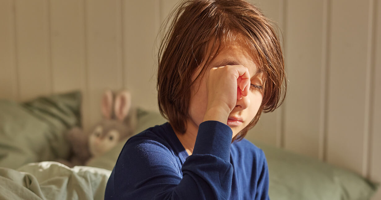 En dreng i blå pyjamas sidder på kanten af sin seng og kløer sit øje, som måske generer ham på grund af husstøvmideallergi